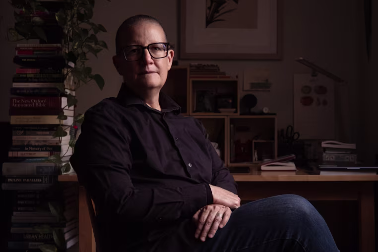 A middle-age white woman with a shaved head sits with crossed legs in front of her desk and large stacks of books.