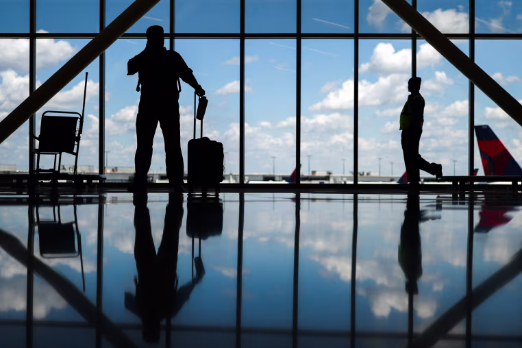 Silhouettes of people standing in an airport near large windows with a view of the sky and tarmac, reflections can be seen on the floor