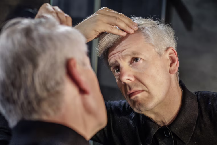 A man examines his hairline in the mirror.