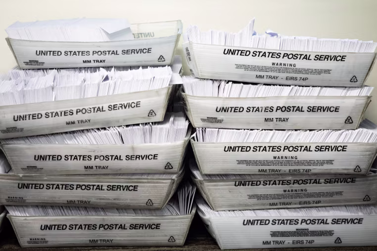 10 United States Postal Service boxes full of envelopes are stacked in two columns of 5 on a table