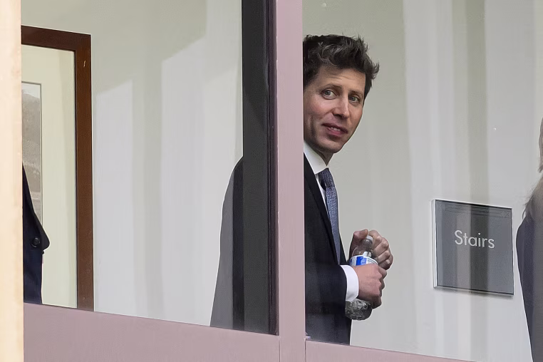 Sam Altman smiles toward the camera through a window. 