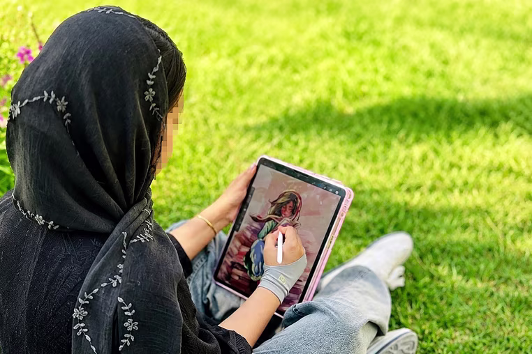 Image: An anonymous photo of a woman drawing a girl on her iPad with a stylus pen. The woman is wearing a black hijab with embroidered flowers.