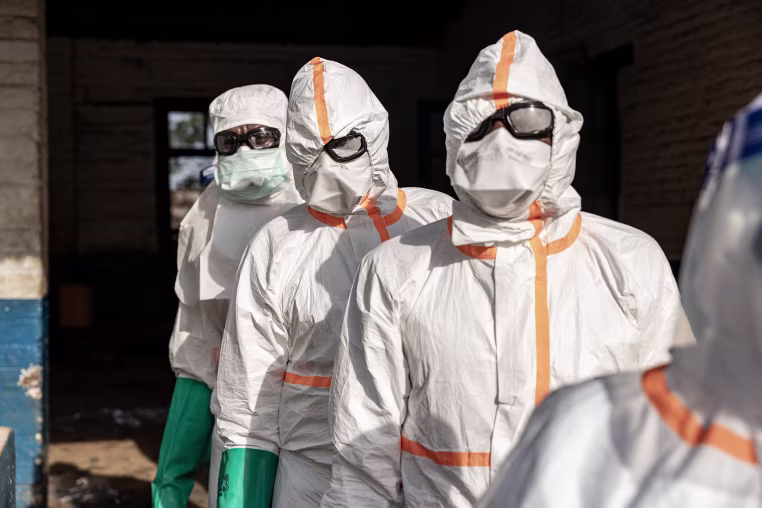 Several medical workers in white hazmat coveralls with masks and goggles standing in a line outdoors.