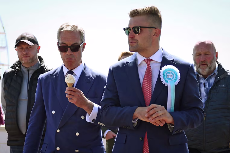 Reform UK leader Nigel Farage eats an ice cream cone as he walks walks with Dan Jukes