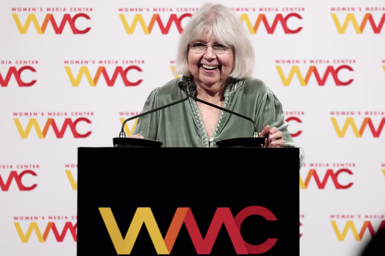 Robin Morgan smiles while standing in front of two microphones on a lectern.