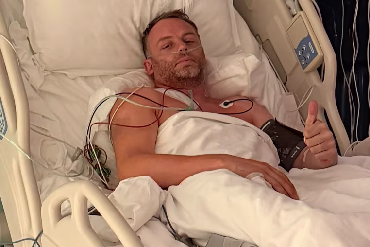 Image: Ollie lying in a hospital giving a thumbs up to the camera