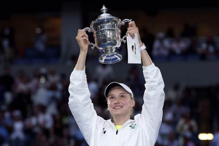 Elena Rybakina holds a silver trophy in the air with both hands.