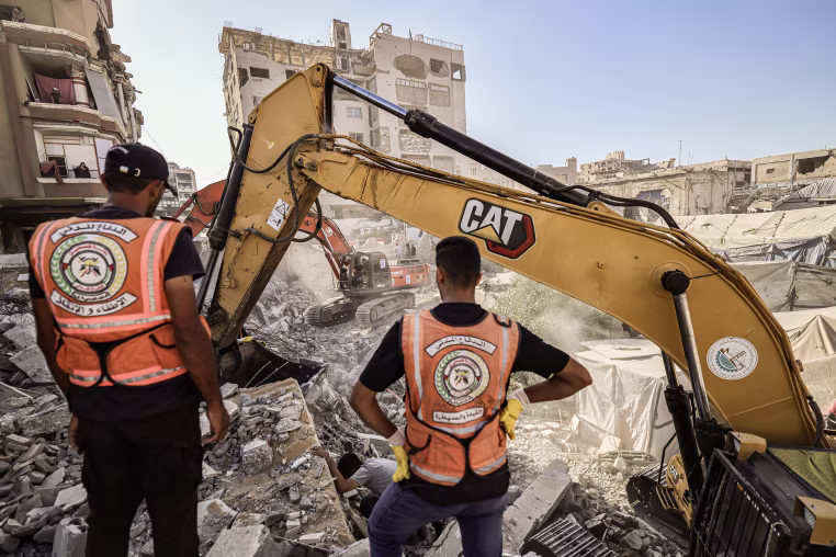 Two men wearing orange vests stand in front of two diggers in a field of debris.