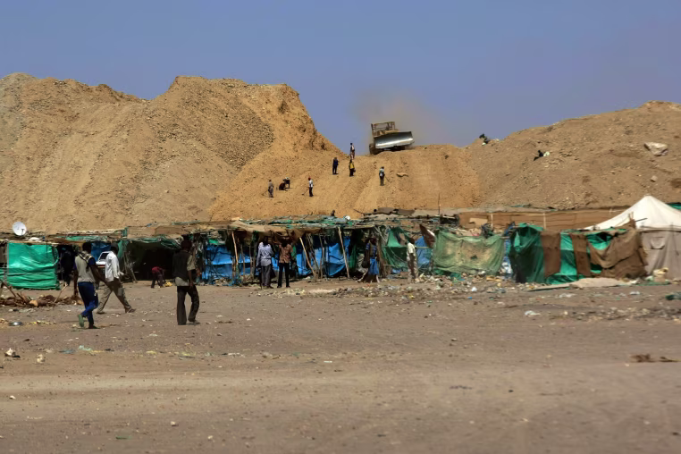 Makeshift shelters lined up against a dry, dusty hillside with a front loader atop it.