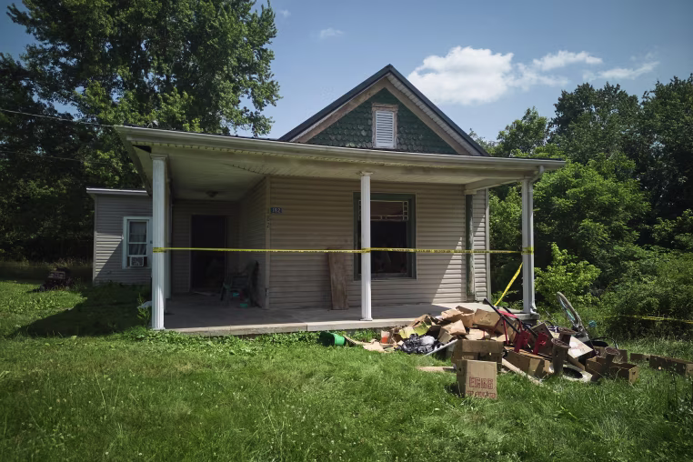 Yellow police tape stretched across the front porch of a small one-story home with a pile of boxes and belongings lying in front of it.