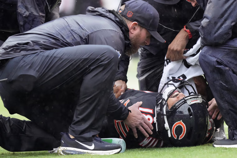 Caleb Williams lying on his back on the field, surrounded by Bears staff.