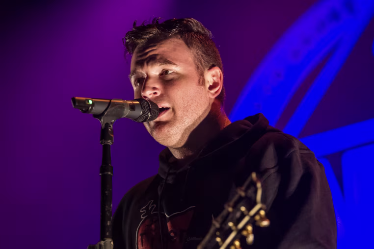 Chad Gilbert singing into a microphone under stage lighting.