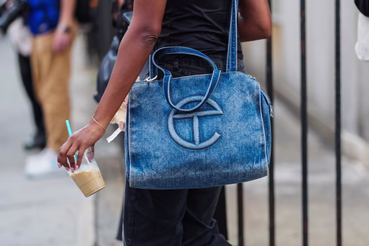 A guest wears a thin gold bracelet on the wrist, a large rectangular washed blue denim Telfar tote with dual top handles, a long shoulder strap, holding a clear plastic cup of iced coffee with a turquoise straw.
