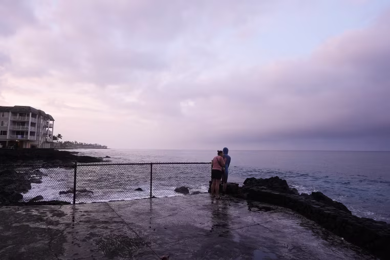 A couple observing a purple sunset over a cloudy sky on the Hawaiian coastline.