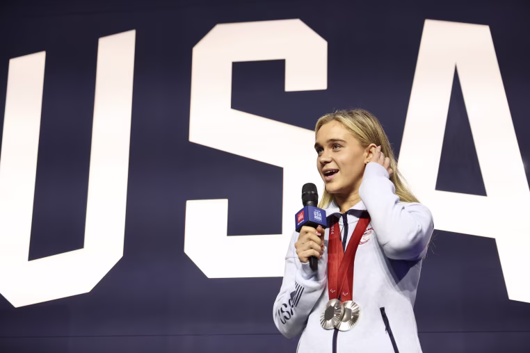 Alexandra Truwit speaking on a stage with two Olympic medals around her neck.