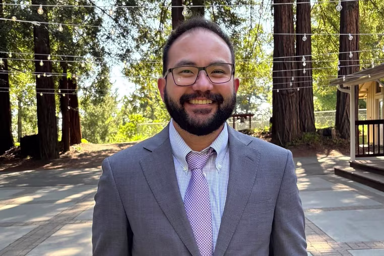 Image: Jonathan McKinsey wearing a suit, stood in front of trees draped with string lights. 