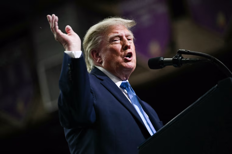 Donald Trump speaks at a podium, lifts one hand up in the air