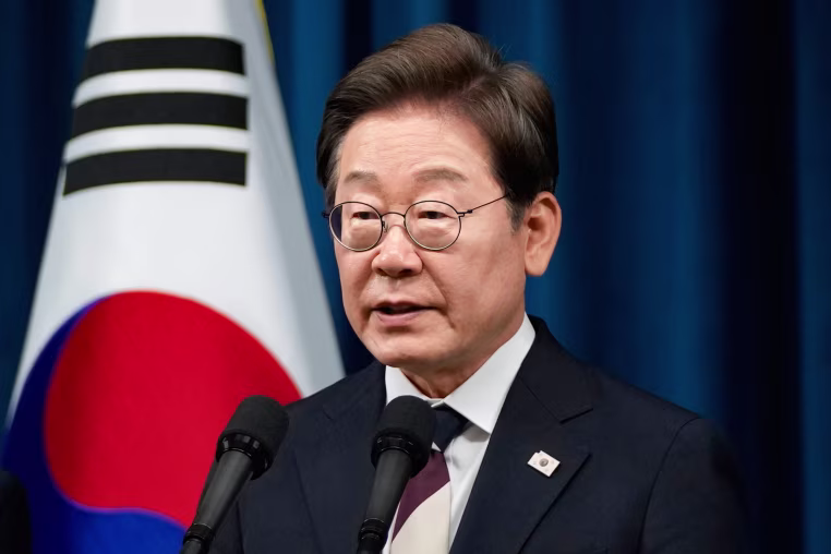 Lee Jae Myung speaks into a microphone, Korean flag can be seen behind him against a blue curtain
