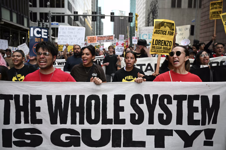 Protestors holding a large white banner reading THE WHOLE SYSTEM IS GUILTY!