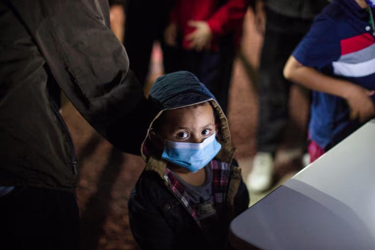A child looks up at the camera wearing a blue facemask while standing outside