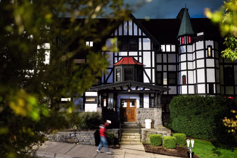 The exterior of the Chi Phi fraternity house, a blurred figure in motion walks by on the sidewalk out front