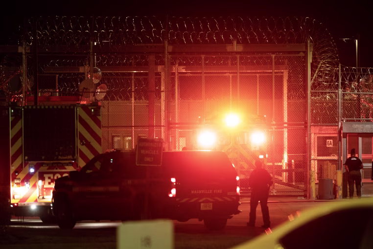 Red lights illuminate the entrance to a prison with tall fencing surrounded by razor wire.