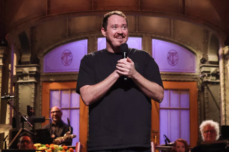 Shane Gillis speaks into a microphone on the SNL stage