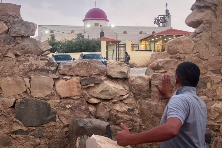 A man looks through a gap in a stone wall toward a Greek Orthodox church.