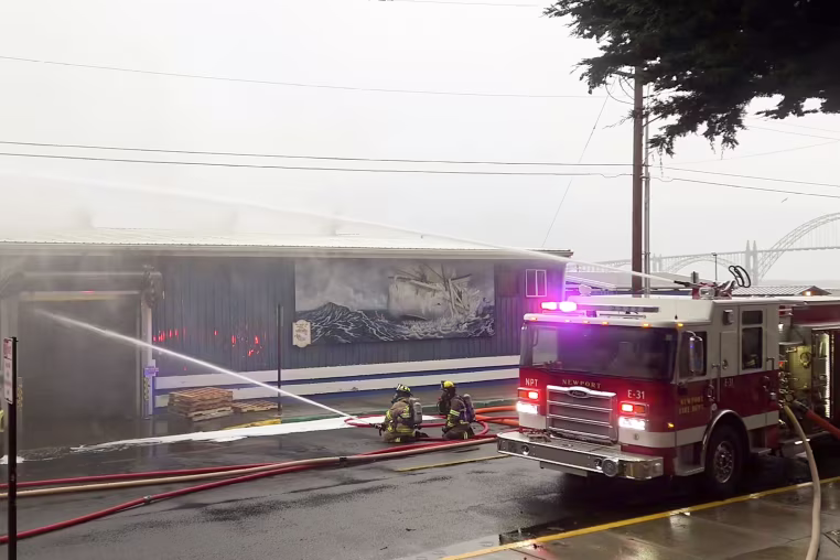 Two firefighters kneel in the street while spraying water on a burning building.
