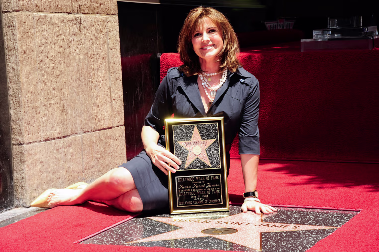 Susan Saint James sits outside on the ground next to her star on the Hollywood Walk of Fame, holding a plaque with a star