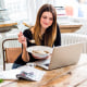 Young woman in city apartment eating muesli breakfast whilst reading laptop