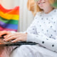 Young Millennial Hippie Woman Sitting On Balcony Using Laptop. Lgbtq Rainbow Flag On Background.