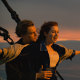 Iconic titanic scene starring Leonardo DiCaprio and Kate Winslet. DiCaprio stands behind Winslet, his hands on her waist and she has her arms outstretched as they look out over the water from the bow of a ship.