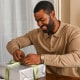 man in a tan sweater opening a gift in silver wrapping paper and a green ribbon.