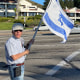 A man waves an Israeli flag in a park space along a city street
