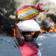 A woman carrying two bags of rice walks past burning tires
