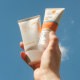 A hand holds two bottles of sunscreen against a blue sky with clouds