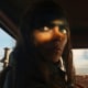 Film still of Anya Taylor-Joy in "Furiosa: A Mad Max Saga." Her face is mostly in shadow and her eyes are highlighted. Trucks are burning in the background.