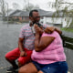 Two people embrace while sitting on a bench as a rain shower soaks them outside, as a volunteer evacuation truck is in the distance