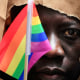 A man covers his face with a paper bag and holds a pride flag in front of his eye