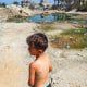 A boy with a skin rash on his back stands near rubble and sewage water