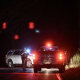 An armed police officer stands on the road with emergency vehicles