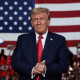 Donald Trump claps with an audience holding "47" signs behind him. A large American flag is the backdrop.