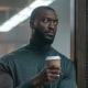 Aldis Hodge leans on a door frame in an office and holds a coffee