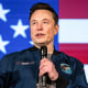 Elon Musk speaks on stage, with an American flag backdrop behind him