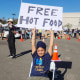 A boy sits in a chair and holds up a sign that says "Free Hot Food" outside