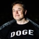 Elon Musk smiles and opens up his jacket to show a black shirt that reads "DOGE" in white lettering