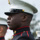 Marines stand outside in uniform and hold rifles, American flag flies in the background