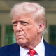 Donald Trump stands outside behind a shrub as a marine in the background salutes him