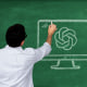 Teacher looking at a chalkboard, drawing a computer with the Chat GPT logo in chalk.